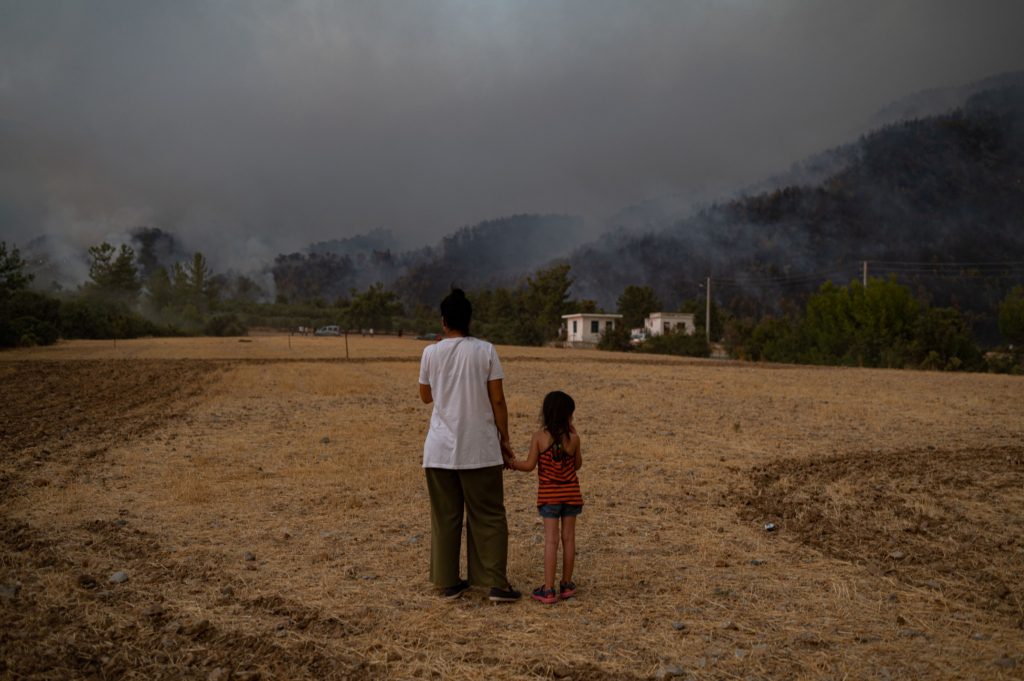 一名妇女和一名儿童站在干燥的沙路上，他们的脸从镜头前移开，看着黑暗、朦胧并充满野火所产生烟雾的天际线。
